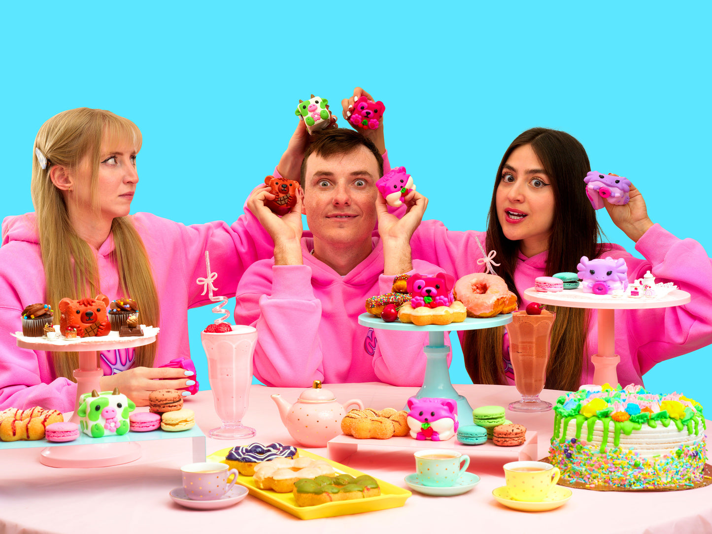 Erin, Christian, and Anna showing their Mushykinz taba squishies while having a tea party with colorful desserts and toys on a blue background